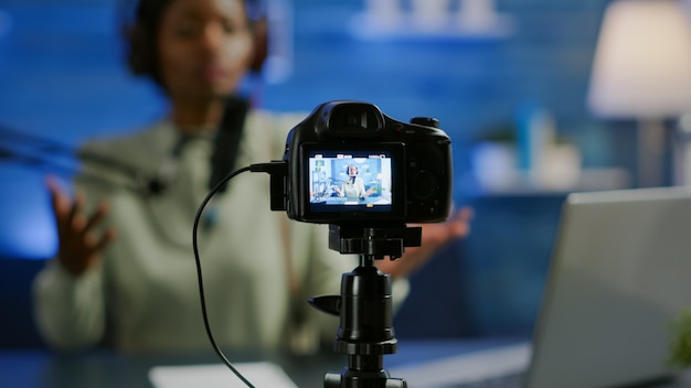 Close up of professional camera sitting in front of dslr recording online show. African woman vlogger speaking at microphone during livestreaming, blogger discussing in podcast wearing headphones