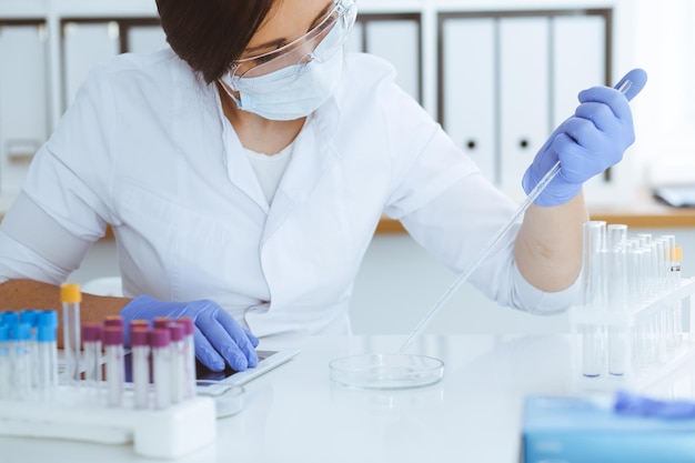 Close-up of professional female scientist in protective eyeglasses making experiment with reagents or blood test in laboratory. Medicine, biotechnology and research concept.