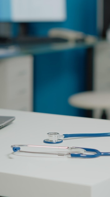 Close up of professional stethoscope placed on white desk in empty doctors office at facility. Medical tool and laptop on table with nobody in cabinet. Healthcare equipment for examinations