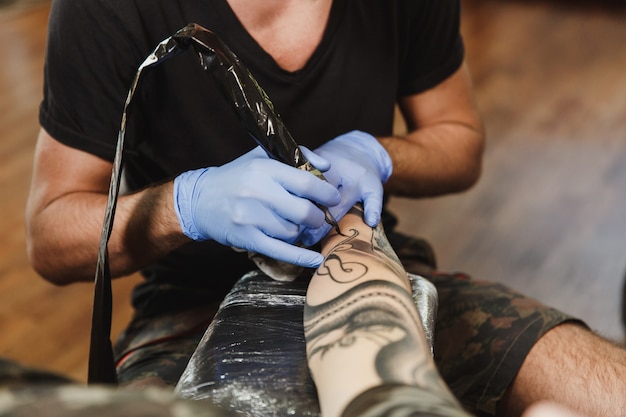 Close up of professional tattooer artist doing tattoo on the arm of a young man by machine with black ink