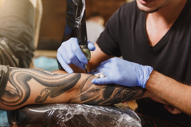 Close up of professional tattooer artist doing tattoo on the arm of a young man by machine with black ink