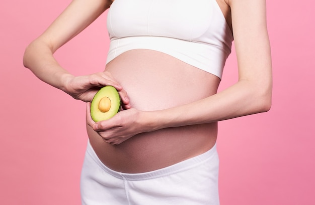 Close-up, profile of a young Caucasian woman, with beautiful healthy skin, in a white top, holding a half avocado at belly level with two hands, on a pink background. Pregnancy and nutrition concept.