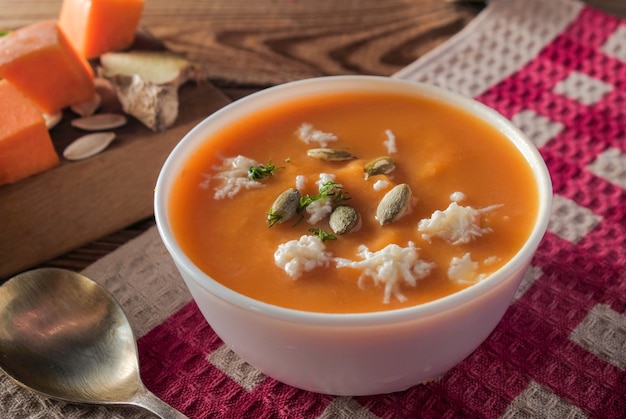 Close-up Pumpkin spicy cream soup with cheese and seeds on a wooden table on a red tablecloth.