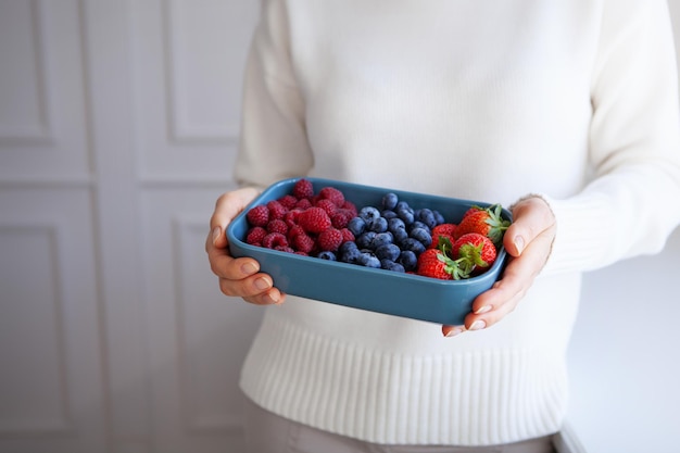 Close-up of raspberries, strawberries, blueberries in blue bowl in hands of woman in white sweater at light background. Copy space and Depth of field