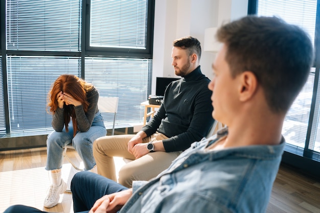 Close-up of sad crying young woman sharing problem sitting in circle during group therapy session. depressed redhead female telling about mental problem to other patients at office.