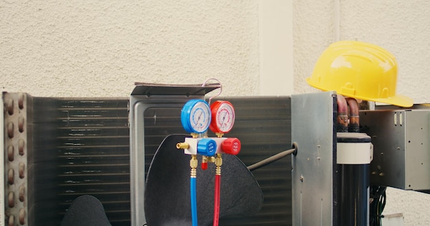 Close up of safety hardhat and pressure measurement device used for checking HVAC system refrigerant in need of maintenance. Set of freon levels benchmarking tool mounted on air conditioner