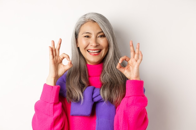 Close-up of satisfied asian female with grey hair, smiling pleased and showing OK signs, approve and like product, praising something awesome, white background.