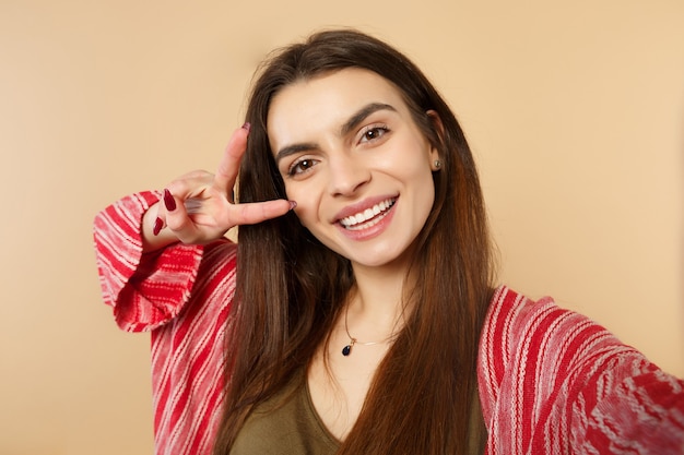 Close up selfie shot of cheerful young woman in casual clothes looking camera, showing victory sign isolated on pastel beige background. People sincere emotions, lifestyle concept. Mock up copy space.