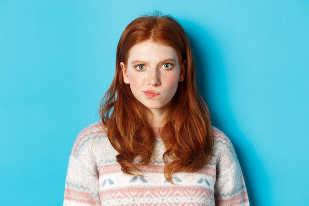 Close-up of serious-looking girl thinking, staring at camera troubled and grimacing, making hard choice, standing over blue background.