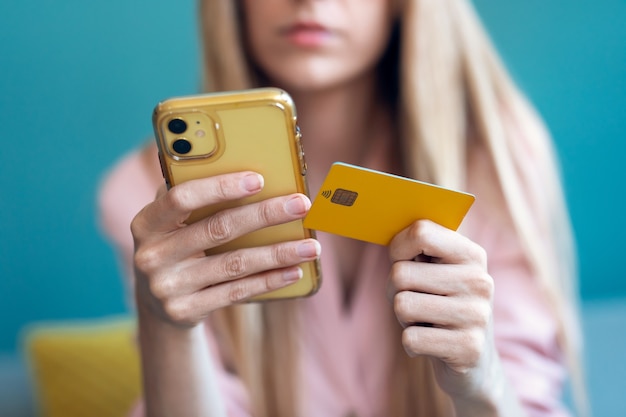 Close-up of serious young woman paying something online with her credit card and the smartphone while sitting on sofa at home.