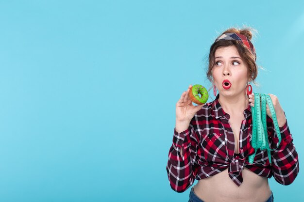 Close-up shocked surprised young beautiful woman looking at measuring tape and holding donut in hands posing on blue background with copy space. Diet and rejection of harmful high-calorie foods.