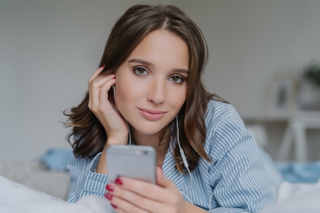 Close up shot of attractive brunette young woman downloads songs from network uses smart phone for entertainment dressed in nightclothes rests in bed has lazy day looks seriously at camera