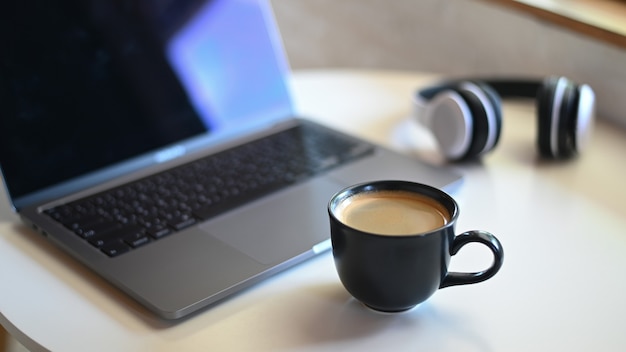 Photo close-up shot of coffee mug with blurred laptop and headphone on placed on a table in a cafe.