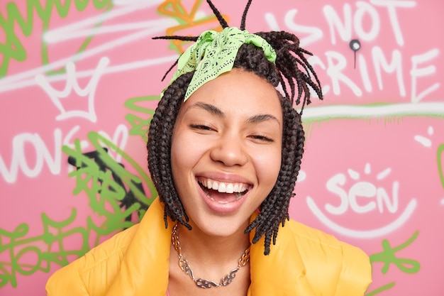 Close up shot of ethnic teenage girl smiles broadly dressed in street style has tendy dreadlocks hairstyle poses against graffiti wall being part of youth gang