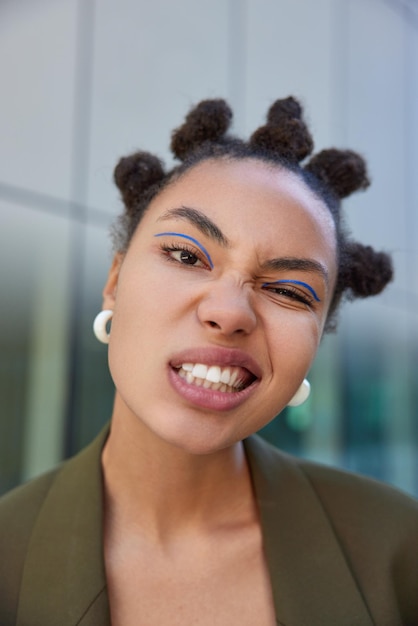 Close up shot of fashionable young woman with bun hairstyle has cheeky expression clenches teeth wears earrings and dark green jacket poses against blurred background has bright vivid makeup.