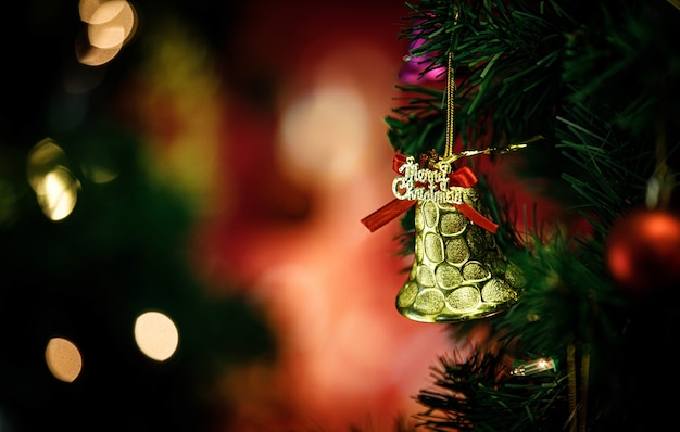 Close up shot of golden glossy glitter bell with red ribbon hanging decorating on green Christmas pine tree branch in front of blurred background in Xmas eve traditional festival celebration night.