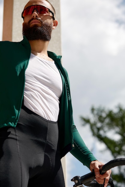 Close up shot of happy attractive young European man with beard wearing glasses and green sportswear looking aside during evening ride in park on weekend