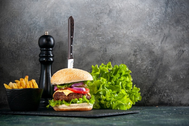 Close up shot of knife in delicious meat sandwich and green fries on black tray on gray surface