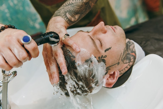 Close up shot of man getting trendy haircut at barber shop. The male hairstylist in tattoos serving client, washing head