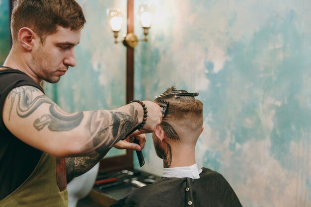 Close up shot of man getting trendy haircut at barber shop. The male hairstylist in tattoos serving client.