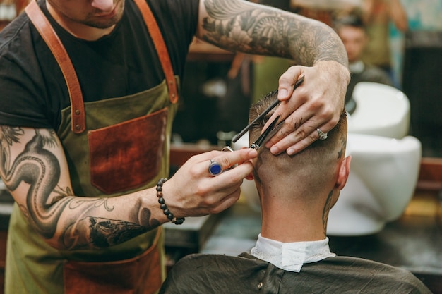 Close up shot of man getting trendy haircut at barber shop. The male hairstylist in tattoos serving client.