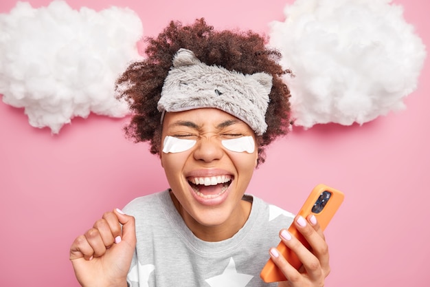 Close up shot of overjoyed ethnic woman with curly Afro hair smiles broadly raises hand feels very happy wears pajama uses mobile phone for surfing social networks poses indoor after awakening