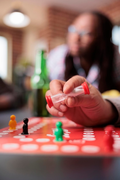 Close up shot of playful man holding boardgame hourglass in hand while sitting at table. Person having hourglass while sitting at home in living room with multiethnic friends while playing table games