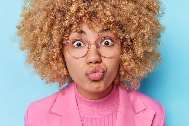 Close up shot of surprised young woman with curly hair focused wondered keeps lips rounded wears transparent eyeglasses and formal clothes isolated over blue background Human facial expressions