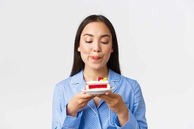 Close-up shot of thoughtful cute asian girl standing at night in pajama with plate of cake