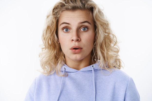 Close-up shot of wondered and surprised cute european female with blue eyes and blond curly hair raise eyebrows in amazement open mouth from astonishment, impressed over white wall