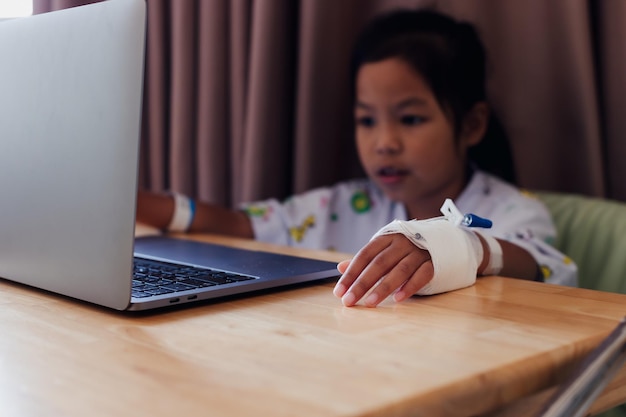 Close up on sick child's hand who have IV solution while child studying online video call with laptop computer in the hospital. Isolation homeschooling and distance learning.