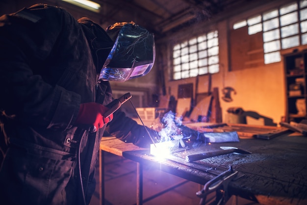 Close up side view of focused professional welder with protection mask working with metal and sparks in a fabric workshop