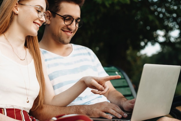 Close up side view of a womans hand pointing on a screen of a laptop smiling while her boyfriend is holding laptop and looking laughing outside in the park.