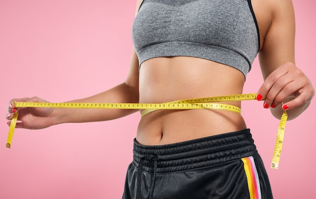 Close up of slim woman measuring her waist's size with tape measure. Isolated on pink background.