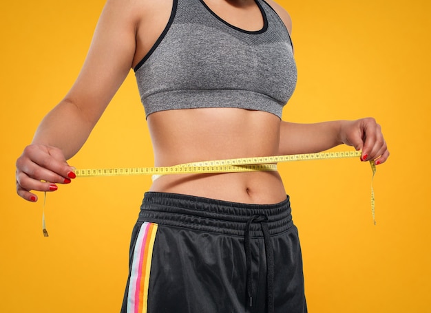 Close up of slim woman measuring her waist's size with tape measure. Isolated on yellow background.