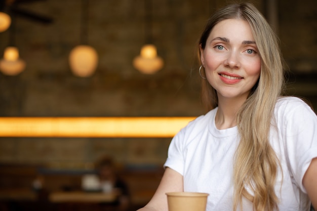 Close-up of a smiling blonde woman in a white t-shirt who is sitting in a cafe with a paper cup. place for your design