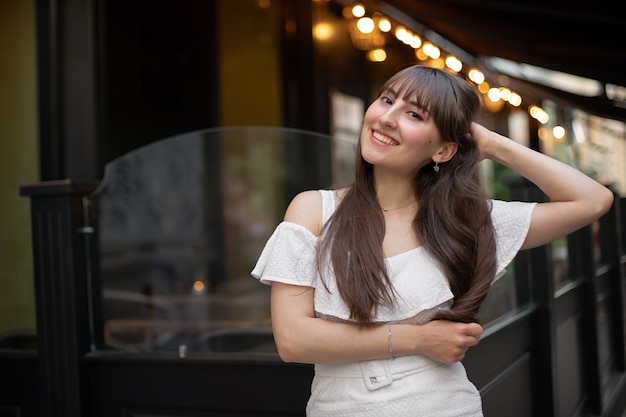 Close-up of a smiling brunette woman with long hair in a white dress on the background of a cafe and yellow lanterns. place for your design