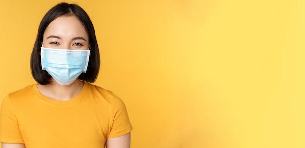 Close up of smiling happy asian woman wearing medical face mask from covid standing in yellow tshirt...