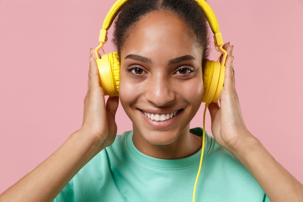 Close up of smiling young african american woman girl in green sweatshirt posing isolated on pastel pink wall. People lifestyle concept.  Listen music with headphones.