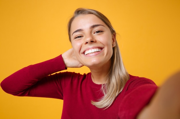 Close up of smiling young blonde woman girl in casual clothes posing isolated on yellow orange background in studio. People lifestyle concept. Mock up copy space. Doing selfie shot on mobile phone.