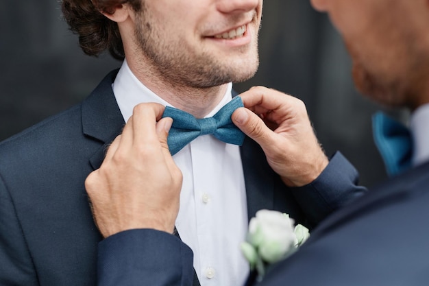 Close up of smiling young man fixing groom bowtie during wedding ceremony same sex marriage concept