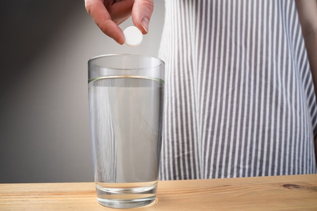 Close up sparkling water glass with dissolving effervescent aspirin pill standing on wooden table, sick unhealthy woman taking emergency medicine, painkiller to relieve headache or fever