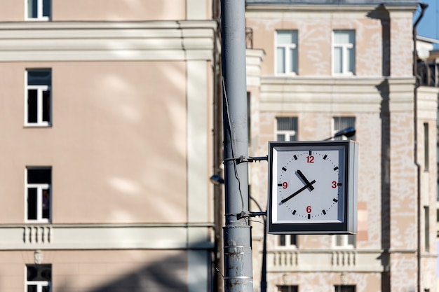 Close-up of a square street clock hanging on a pillar against the background of a brick wall of a building