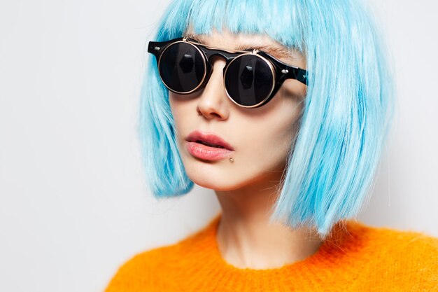 Close-up studio portrait of pretty girl with blue hair, wearing round black sunglasses and orange shirt on white background. Fashion concept.