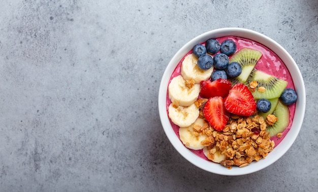 Close-up of summer acai smoothie bowls with strawberries, banana, blueberries, kiwi fruit and granola on gray concrete background. Breakfast bowl with fruit and cereal, top view, space for text