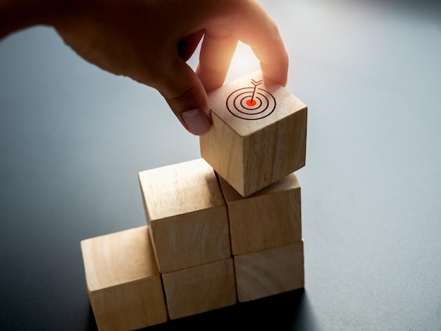 Close Up Target icon on the top of wooden cube block graph chart step put by hand on black background Business achievement goal and objective target investment growth and development concepts