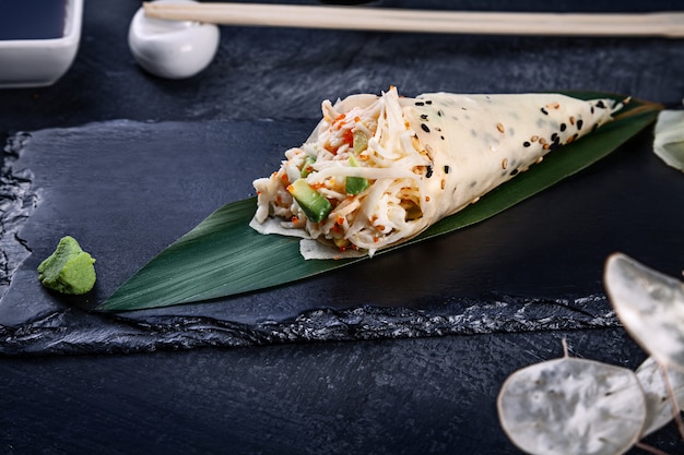 Close-up of tasty hand roll sushi in mamenori with crab and Tobico caviar served on dark stone plate with soy sauce and ginger