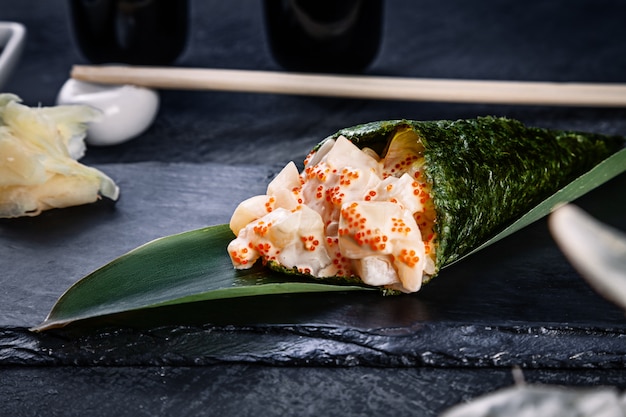 Close-up of tasty hand roll sushi with scallop and Tobico caviar served on dark stone plate with soy sauce and ginger