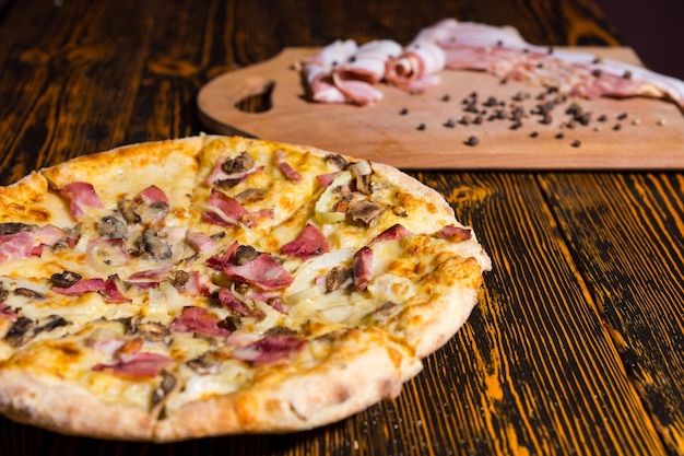 Close up of tasty pizza with variety of toppings and cheese on wooden table, near lies cutting board with bacon and spices