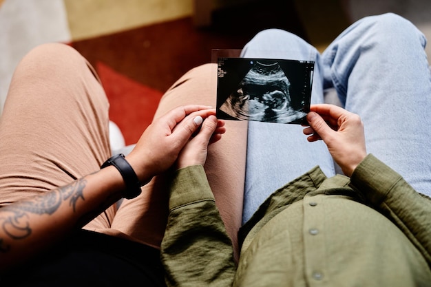 Close up of tattooed gay couple with pregnant young woman looking at ultrasound image of baby same s